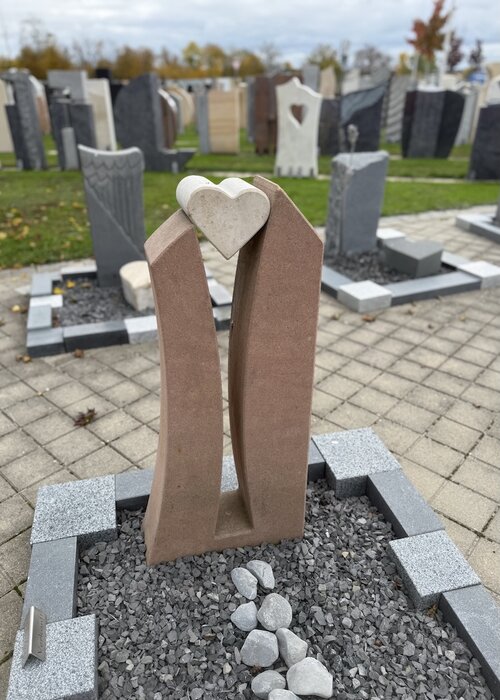 Ein Grabstein für ein Urnengrab angefertigt aus rötlichem Sandstein. Zwei emporsteigende Stelen nehmen ein herzförmigen Kalkstein auf. Trotz der schmalen Flächen bietet der Stein genügend Platz für anzubringende Schriften. | © Messerschmidt GmbH