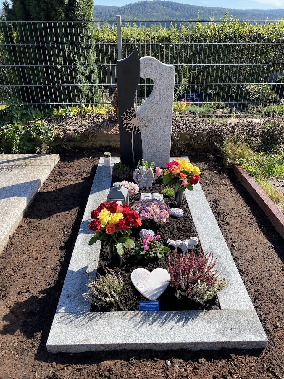 Grabstein Modell Lyon aus Granit und Basalt mit passender Einfassung aus hellgrauem Granit. Mit davor gestelltem Edelstahlornament mit dem Motiv einer Pusteblume. | © Messerschmidt GmbH